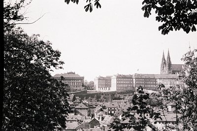 Panoramic view of a European cityscape, likely Prague. Features layered rooftops, stately institutional buildings, and a prom...