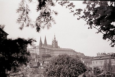 A view of Prague Castle, seen through foliage. Buildings and rooftops fill the foreground, leading to the castle complex with...