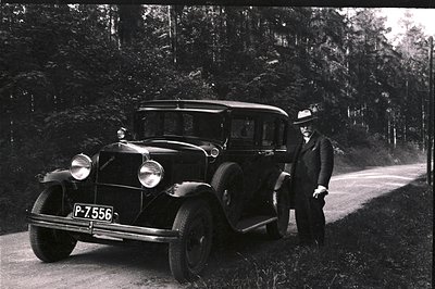 Vintage black sedan, likely a 1920s Chevrolet, stands on a rural road. A formally dressed man with a hat poses beside the car...
