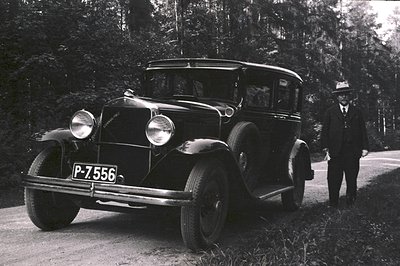 Classic 1930s sedan, possibly a LaSalle, sits on a gravel road flanked by trees. A formally dressed man with a hat stands bes...