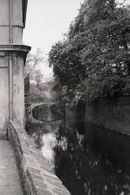 A long, narrow canal is framed by a stone wall and dense foliage. A bridge reflects in the still water, creating a symmetrica...