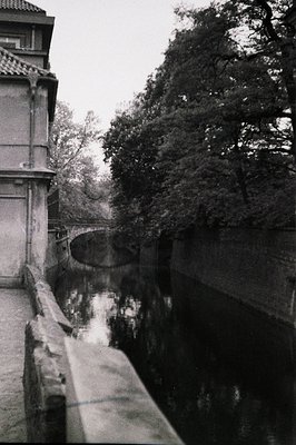 High-angle view of a canal, framed by a weathered stone balustrade and dense foliage. Likely a European canal scene, potentia...