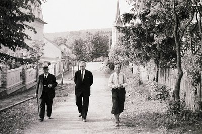 Three men in suits walk along a gravel road lined by overgrown foliage and a weathered wooden fence. A church steeple rises i...