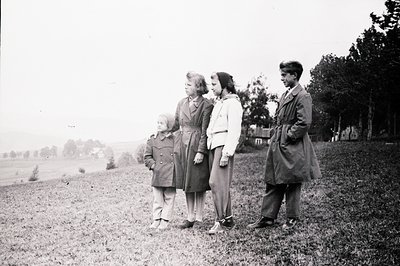 Four children, likely siblings, stand on a grassy slope overlooking a landscape of scattered buildings and trees. All wear tr...
