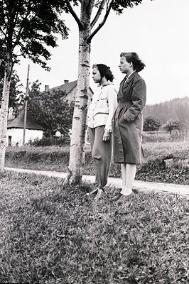 Two young women stand beside a birch tree, dressed in 1940s-style attire. One wears trousers and a light jacket; the other, a...