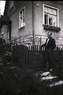 A man stands near a picket fence in front of a modest, stucco-covered house. He wears work clothes and boots, appearing to be...