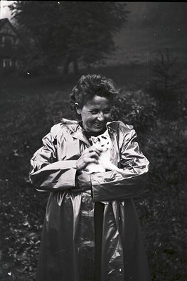 A woman in a light-colored raincoat smiles while holding a small dog or puppy. Natural foliage fills the background, suggesti...