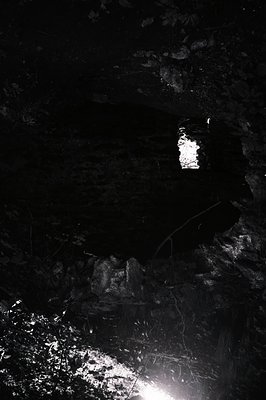 High-contrast monochrome image depicts a small, arched window set within weathered stone. The window reveals a bright, undefi...
