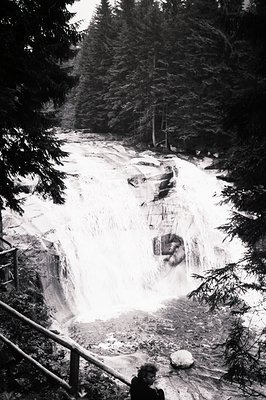 Dramatic, high-contrast view of a powerful waterfall cascading through a rocky gorge. A person stands on a viewing platform o...