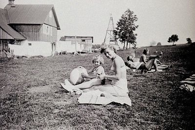 A group relaxes on a grassy lawn near a barn and a wooden water tower, likely a rural farm scene. A woman sits with a young g...