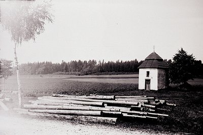 A small, stone chapel with a cross-topped roof sits on a gentle rise, viewed across a field and birch forest. Stacked logs ar...