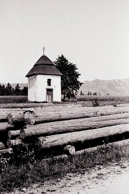 A small, white, Orthodox chapel with a distinctive dome and cross stands before a log barrier. Rural landscape with wooded hi...