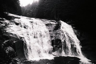 Dramatic, long exposure captures a powerful waterfall cascading over layered rock formations. Dense evergreen forest frames t...