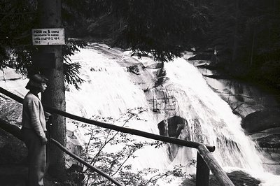 A woman in a hat observes a powerful waterfall cascading over layered rock formations. A warning sign in Cyrillic script is v...