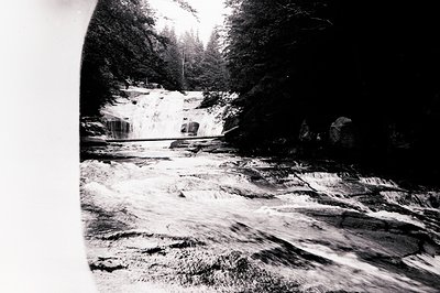 A rugged, forested river cascades over rock formations, creating a series of small waterfalls. Dense trees line the riverbank...
