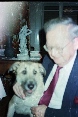 A man in a suit & tie stands alongside a light-colored dog, captured in a vintage-style snapshot. The room features a statue,...