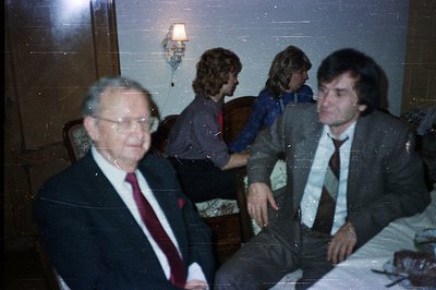 Formal portrait featuring three men. The man on the left wears a dark suit & tie; two men are seated in a patterned armchair....