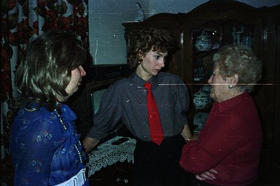 Three individuals stand in a room with a dark floral wallpaper and antique china cabinet. Woman in blue dress, younger woman ...