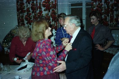 A couple dances closely in a brightly lit interior. The woman wears a patterned red and pink blouse. The man is in a dark sui...