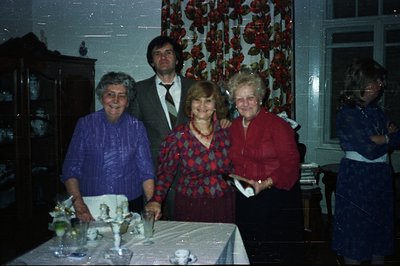 Group portrait of five individuals in an interior space. The décor suggests a 1970s aesthetic, featuring a vibrant floral wal...
