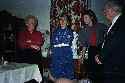 Four people are depicted in a domestic interior, likely a dining room. A man in a suit, a woman in red, and two young women i...