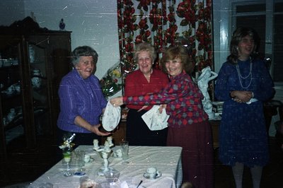 Four women gather around a table laden with decorative objects and a partially obscured tablecloth. Floral wallpaper and stat...