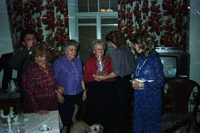 Group portrait within a warmly lit room; six individuals gathered near a table laden with food. Floral wallpaper & vintage te...