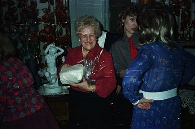 A smiling woman in a red sweater receives a gift wrapped in clear cellophane. Statue and floral wallpaper suggest a domestic ...