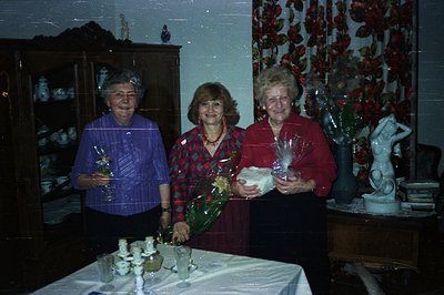 Three women pose indoors, holding floral bouquets. The setting appears to be a dining room with a prominent china cabinet and...