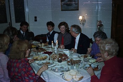 A family gathers around an elaborately set table, laden with food and drinks. The dining room features a decorative wall scon...