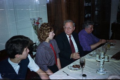 A family gathering at a formal dining table. Four people seated, dressed in 1980s attire. A floral centerpiece sits between t...