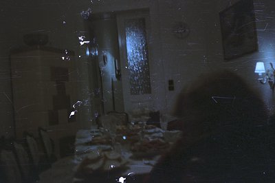 A dimly lit interior shows a table set for a meal, viewed from a shadowed doorway. A portrait hangs on the wall, and a lamp p...