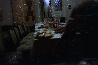 A dimly-lit dining room scene. A long table is set for a meal with visible glassware and food. Visible scratches and dust sug...