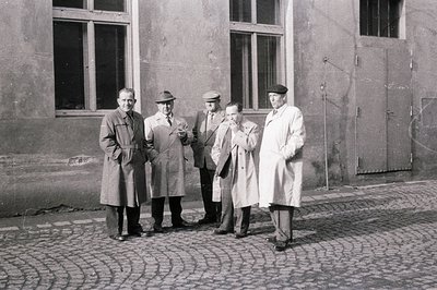 Four men stand outside a building with large windows and metal doors. The men are formally dressed in suits, coats, and a lab...