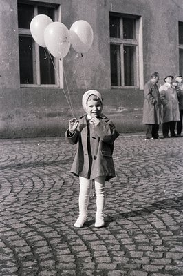 A young girl in a wool coat, stockings, and a headscarf holds three balloons on a cobblestone street. An older group of peopl...