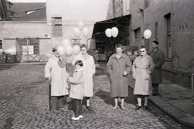 A group of six people, including a child, stand on a cobblestone street amidst a backdrop of industrial buildings. They hold ...