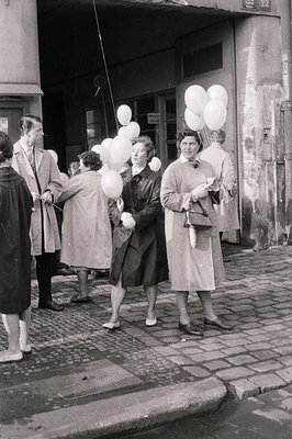 B&W image captures a street scene in what appears to be post-war Europe. Two women hold large bunches of white balloons, alon...