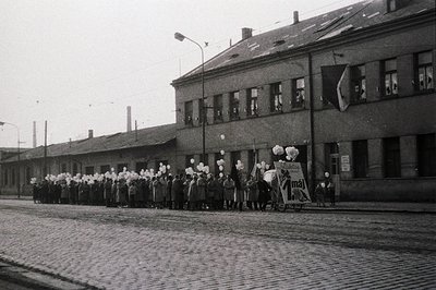 A long procession of uniformed figures, possibly military or paramilitary, marches down a cobbled street lined with a block o...