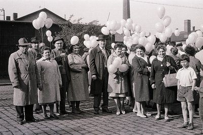 A group of twelve people, mostly women and children, stand amidst a cluster of white balloons on a paved area. Individuals we...