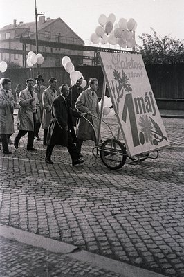 Group of men in overcoats pull a cart bearing a banner reading “1. máj” ("May 1st") and a logo for "Laktos Zdraví" (likely a ...