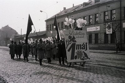 A May Day procession travels along a cobblestone street, featuring a large banner reading "1 máj" (May 1st), along with white...
