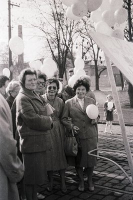 A group of formally dressed women stand amidst a crowd, surrounded by numerous white balloons and decorative fabric. A young ...
