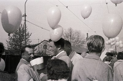 A crowd gathers outdoors, likely for an event or celebration. Numerous large white balloons are suspended overhead, creating ...