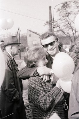 A candid moment captures a couple embracing with a young boy holding a balloon. The man wears a fedora hat & sunglasses, whil...