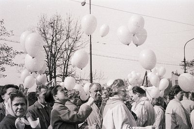 A crowd gathers, releasing a large cluster of white balloons. Men in suits & women in vintage dresses observe. Likely a celeb...