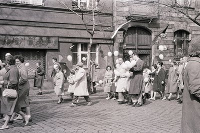 Group of people, many children, strolling down a paved street, holding numerous white balloons. Building facade with arched w...