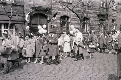 A procession of formally dressed people, many carrying white balloons, walks along a cobblestone street lined with older buil...