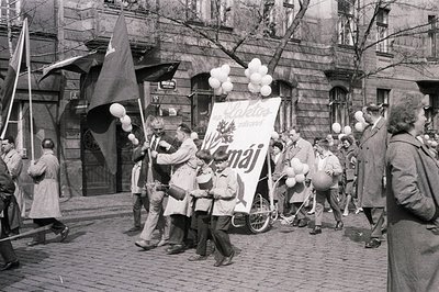 A May Day parade in an Eastern European city. Participants, largely men in overcoats, carry banners with Cyrillic script and ...
