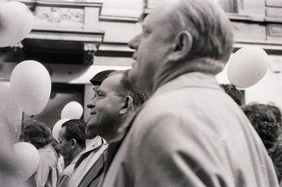 Close-up of two men in profile amidst a crowd, possibly at an event with balloons. One wears a cap, the other a suit jacket, ...