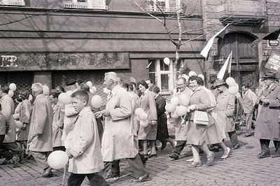 Monochrome image captures a jubilant street procession, likely a celebration or parade. A diverse group of people, mostly adu...
